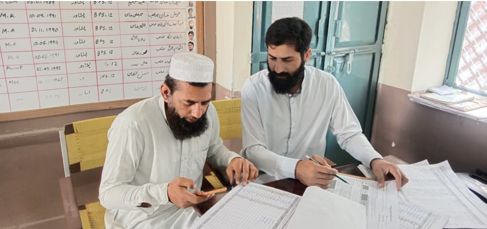 Photo showing 2 teachers performing marking on paper and calculating total scores manually despite having a smartphone.