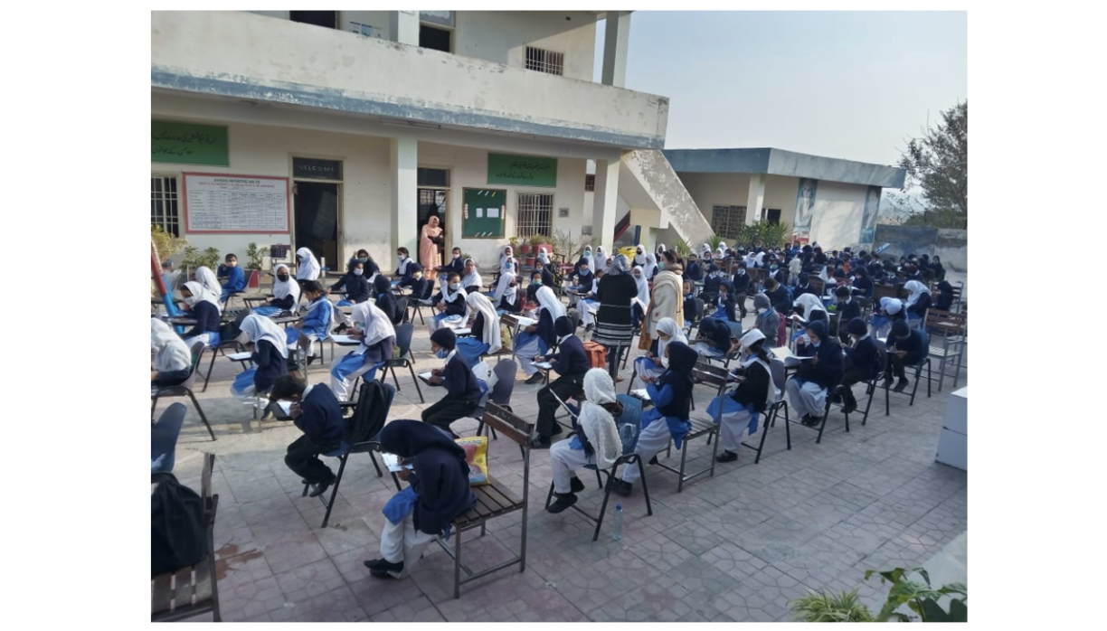 Photo of an outdoor lesson. Pupils are sitting at single desks and there are standing teachers around.